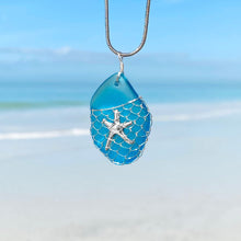 Load image into Gallery viewer, What a Catch Necklace in Ocean Blue hanging close for a shot with a blurred beach background.