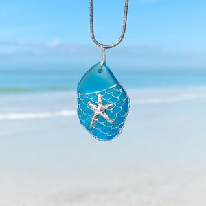 What a Catch Necklace in Ocean Blue hanging close for a shot with a blurred beach background.
